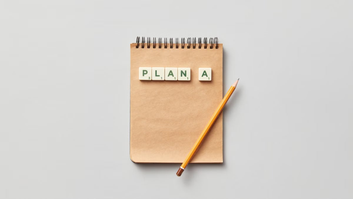 Top view of a notepad with a pencil and scrabble tiles spelling 'PLAN A' on a gray background.